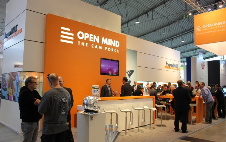 Messestand von OPEN MIND mit Besuchern und Beratern an der Theke. Auf dem Screen an der Wand läuft unser Film.